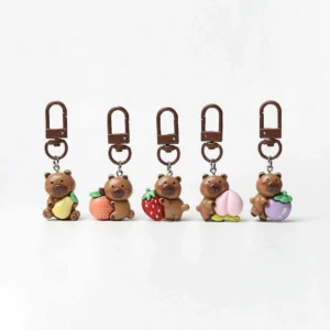 Gesamtes 5-teiliges Capybara Taschenanhänger Obst-Serie Set in einer Reihe angeordnet, jeder Anhänger hält eine andere Frucht: Birne, Persimone, Erdbeere, Pfirsich und Mangostan