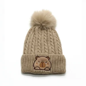 Beige Kinder Capybara Beanie aus Zopfmstrick mit großer flauschiger Bommel und besticktem Capybara-Patch auf dem Umschlag